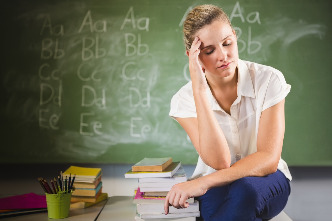 Tensed school teacher sitting with hand on forehead in classroom at school